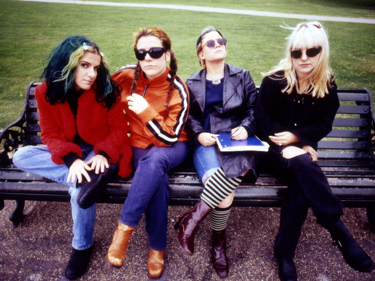 Four women — members of rock band L7 — pose on a park bench wearing sunglasses.