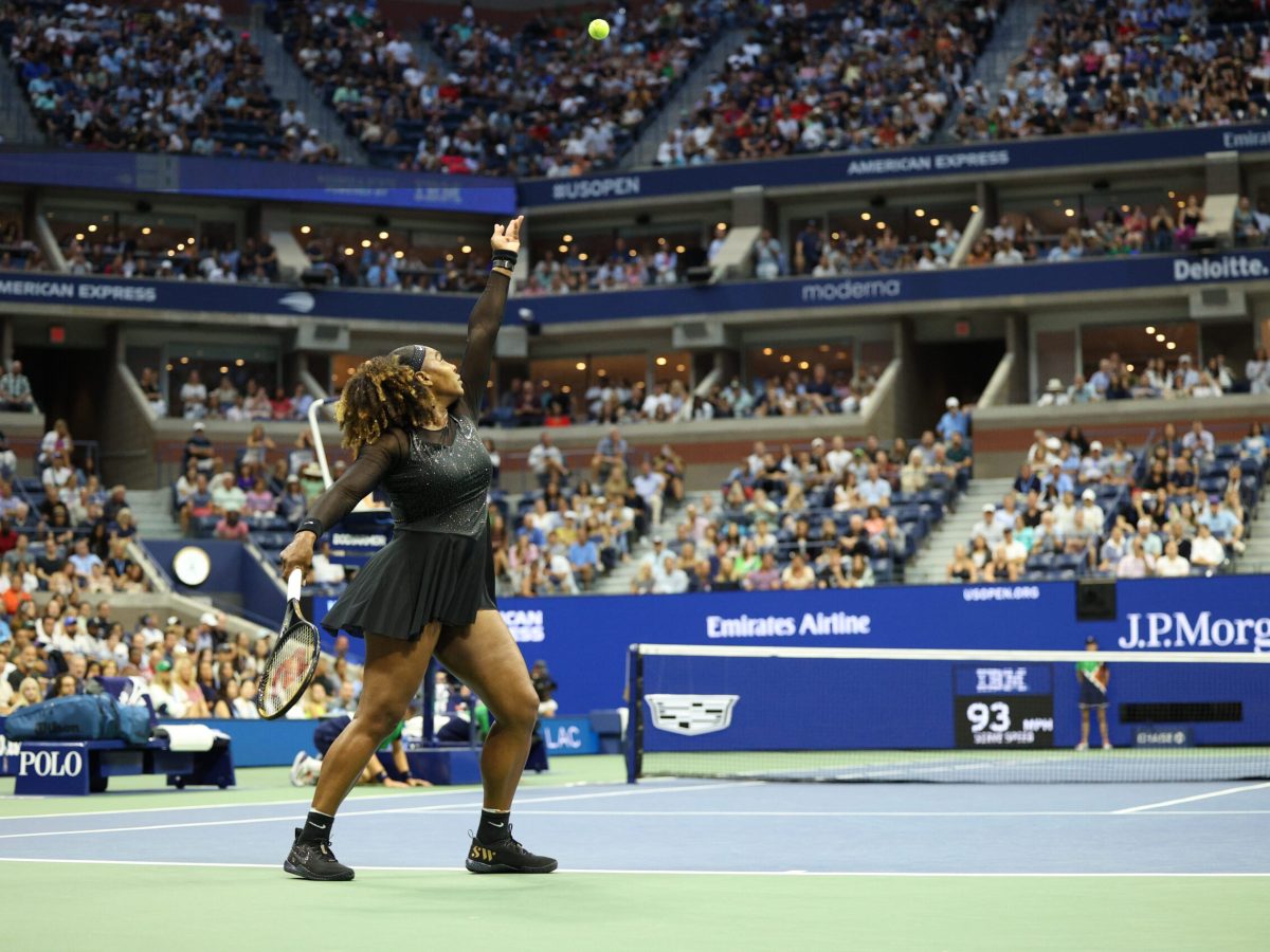 Serena WIlliams, wearing a black tennis minidress, tosses a ball high in the air before serving at the US Open.