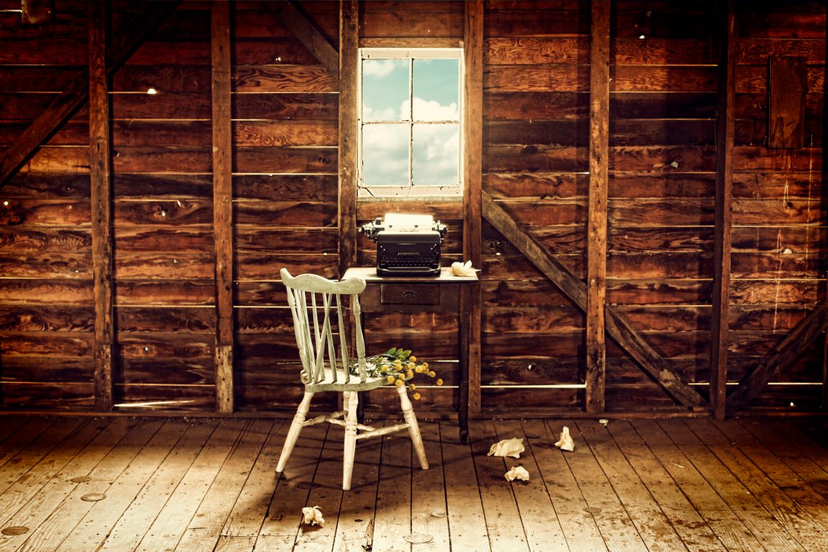An empty chair sits in front of a typewriter and small desk in a desolate wood room with a view.
