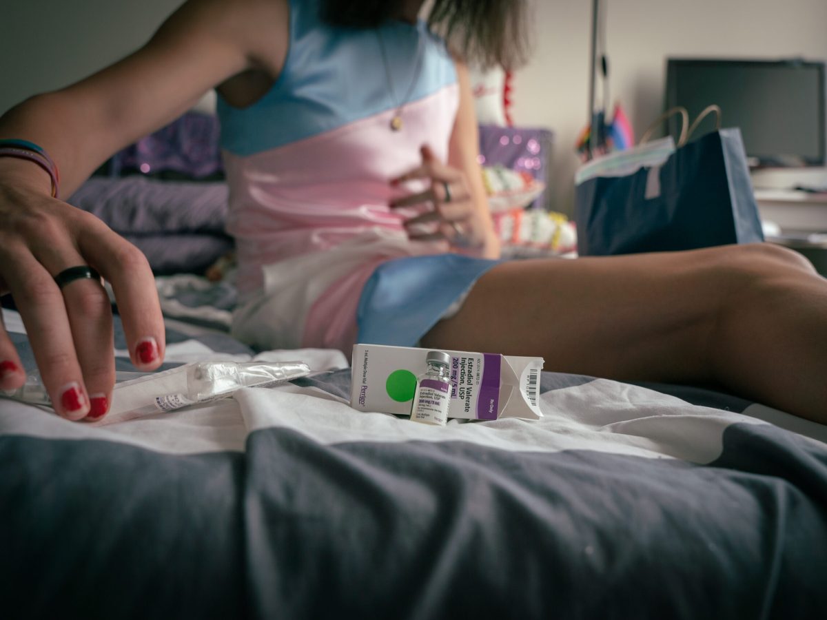 A patient in a blue and pink dress lays out estrogen injection materials on a hotel room bed.