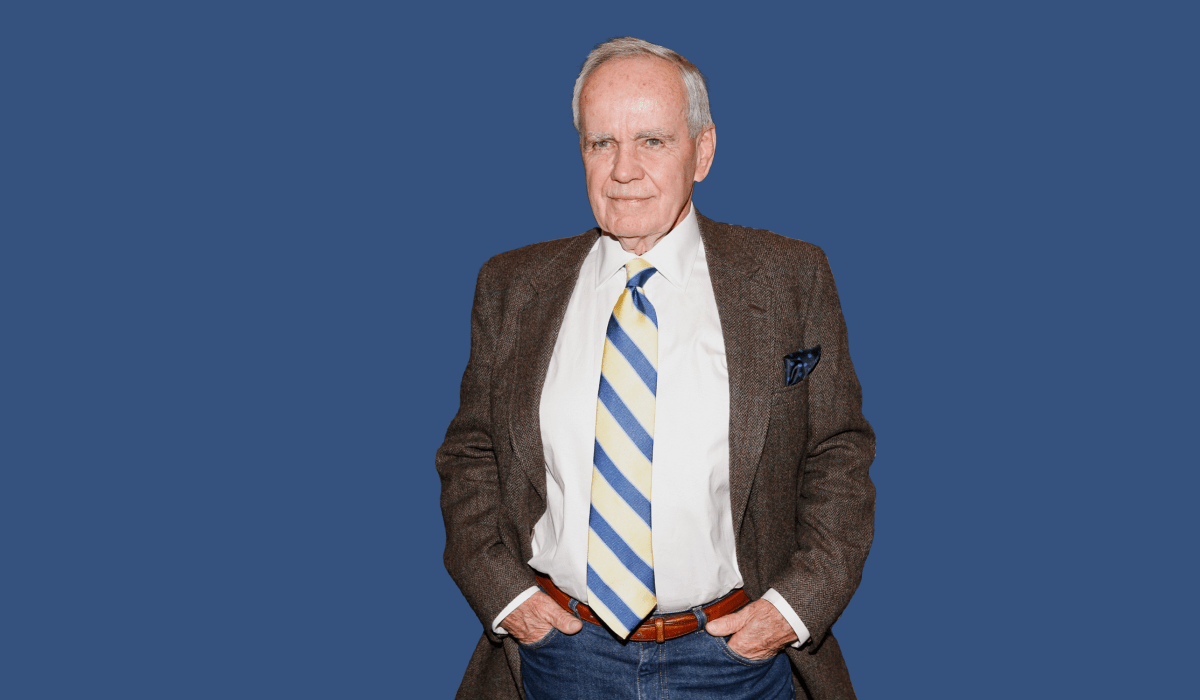 A white man in his seventies—late writer Cormac McCarthy—stands, hands in pocket, against a dark blue background